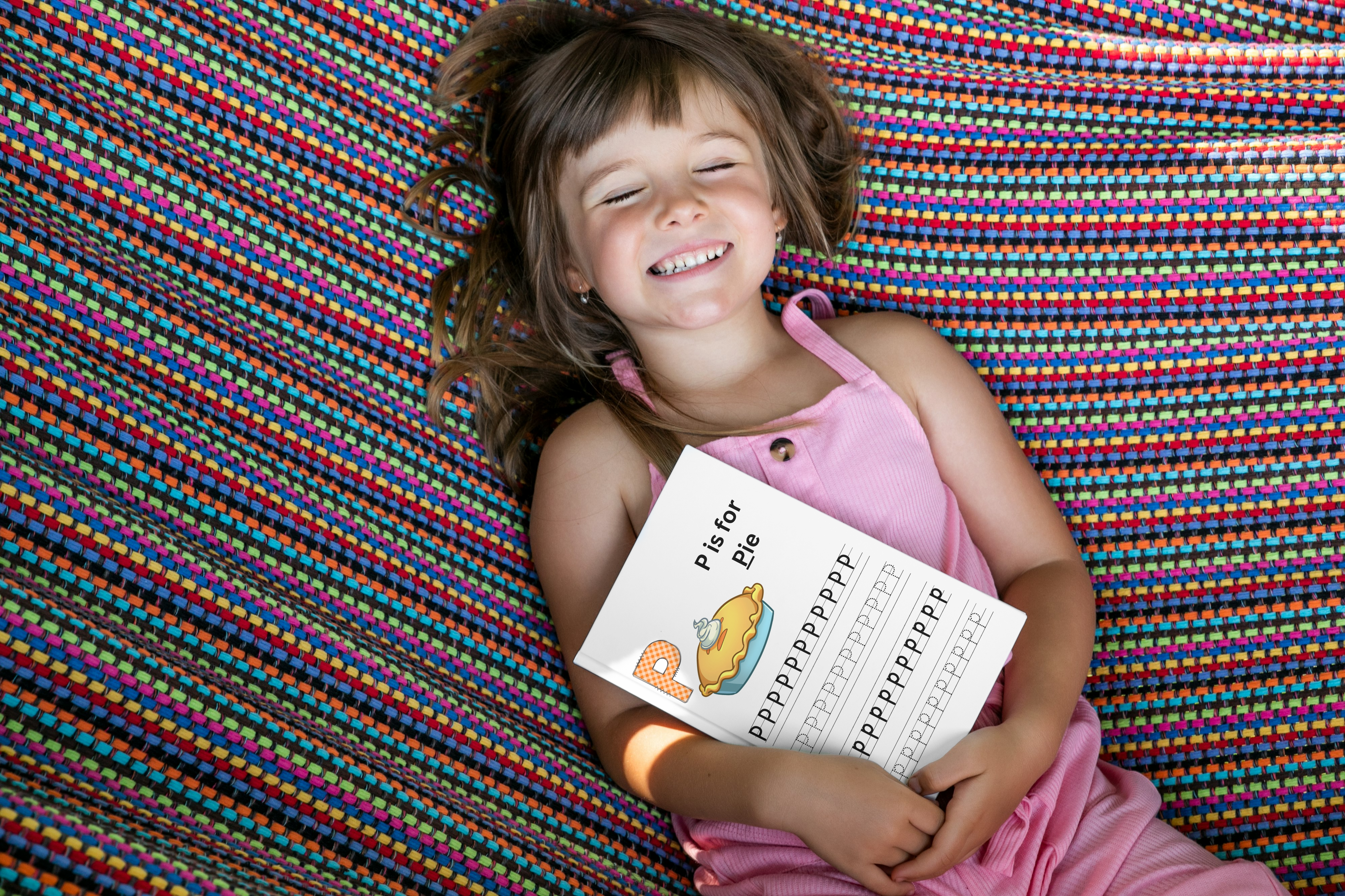 Smiling young girl holding an educational 'P is for Pie' booklet while lying on a colorful hammock, capturing the joy of learning through play and comfort in a vibrant, engaging setting